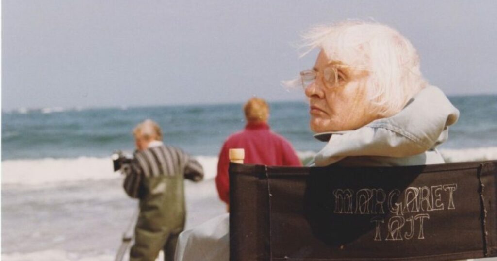 A white woman with white hair and glasses, seated in a director's chair embroidered with the name Margaret Tait, looks over her left shoulder. There is a shoreline, a lively sea with breaking surf and clear horizon behind her, and a cameraperson wearing green waders and a striped sweater is looking out to sea on the beach. A person wearing a red jacket stands next to the cameraperson, also looking out to sea.