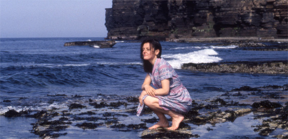 A white woman in a light summer dress crouches on a stony beach, looking out at the waves, with a cliff rising behind her.