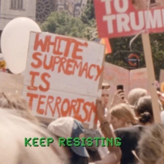 A protest with signs that include White Supremacy is Terrorism and a balloon shaped like Trump's hair. There is a subtitle in green dot-matrix font that reads: Keep resisting.