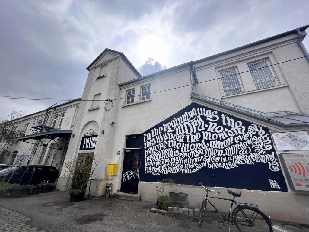 A mural in wave-like white uncial script on a black pentagonal space at the base of a white wall. The text is broken up into diagonal fragments, but reads "In the beginning was the Word. 
No.

This is about the moment before the word, when everything inside you is broken open.

Words, together; multiple and multivalent, coming in a rush, sounding in a chorus, a force like a waterfall. A dam breaking. A difficult birth." 

There is a black door to the left of the wall that has some white graffiti sprayed on it. The main door of the theatre and sign that reads Pathos is just visible past the grafitti'ed door.