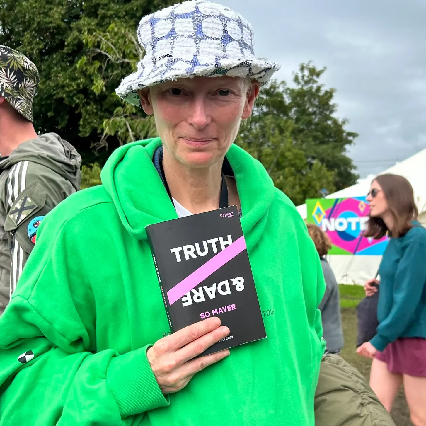 Tilda Swinton holds a copy of the proof of TRUTH & DARE. She is outdoors at a festival, and is wearing a bright green Renaissance tour hoodie and a floral bucket hat.