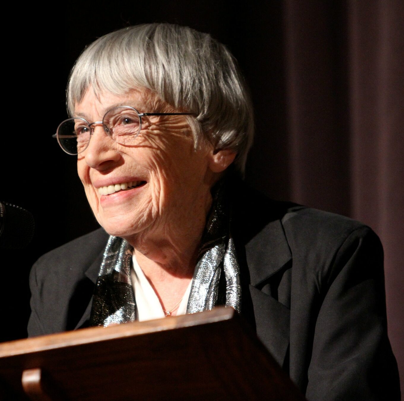 Ursula K Le Guin, an older woman with a white bowl cut and wearing glasses, stands at a lectern, smiling